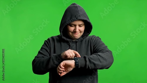Young person in a dark hoodie looking at their smartwatch on a green screen background.