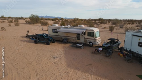Aerial orbit of a desert camp with motorhomes, trailers, and UTVs parked in a circle.