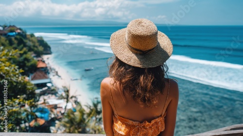A woman sits at a viewpoint overlooking a beach and ocean in Bali. She wears a straw hat and looks at the waves and shoreline. Trees surround the area.
