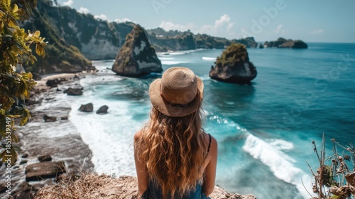 A woman with long hair wearing a hat sits on a cliff edge. She looks out over the ocean waves crashing on the rocks below. The sun shines brightly on a clear day in Bali.