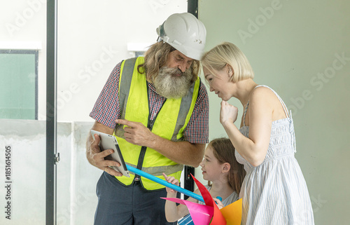 Experienced bearded foreman shows new home plans on a tablet to a smiling mother and daughter, highlighting family trust, smart construction technology and a warm, hopeful future in their house