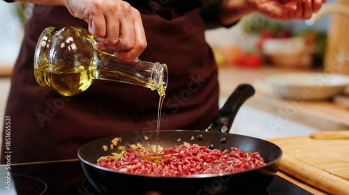 Close-up of chef pouring olive oil into a frying pan with raw ground meat and onions during cooking.