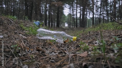 An empty plastic bottle lies discarded on the ground in a pine forest. The single-use waste pollutes the natural environment surrounded by trees and grass.