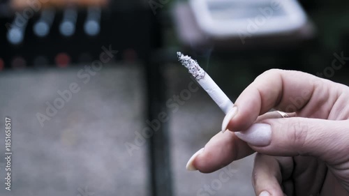 A woman holds a smoking cigarette with manicured fingers in an outdoor setting near a barbecue. The wisps of smoke drift upward from the burning tip highlighting nicotine dependence.