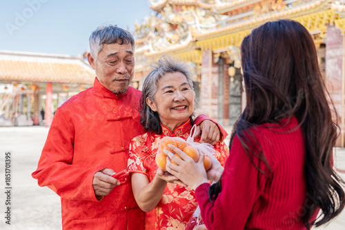 During the Lunar New Year at a beautiful Chinese temple, a cheerful teenage daughter presents her parents with oranges, symbolizing love and respect. Together, they cherish this festive tradition.