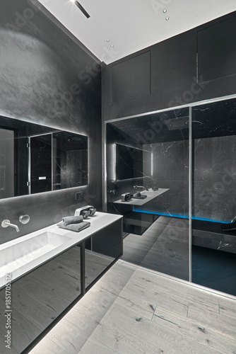 Modern black bathroom interior with parquet flooring, sink cabinet in the left foreground and glass shower enclosure in the background, minimalist contemporary design