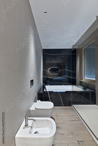 Close-up of white porcelain bidet and toilet in a modern domestic bathroom. Background shows a large bathtub. Hardwood flooring and bright lighting.
