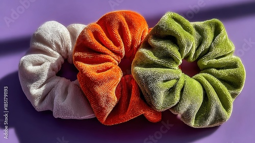 Three scrunchies in white orange and green colors lie on a purple surface in bright sunlight.