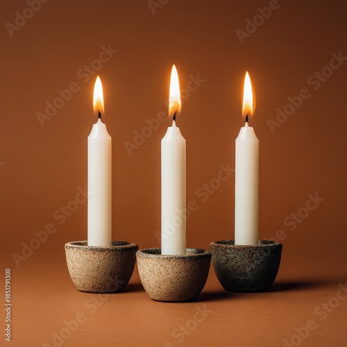 Three white taper candles with lit flames stand in textured candle holders against a warm brown background.