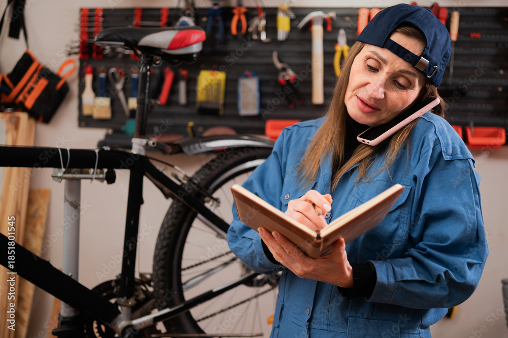 Fototapeta premium Woman bike mechanic phone call with notebook for writing notes work in bicycle repair shop or maintenance garage. Concept of small business, start-up and vehicle workshop