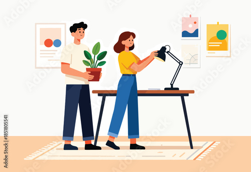 Two people arrange desk with plant & lamp next to artwork on wall