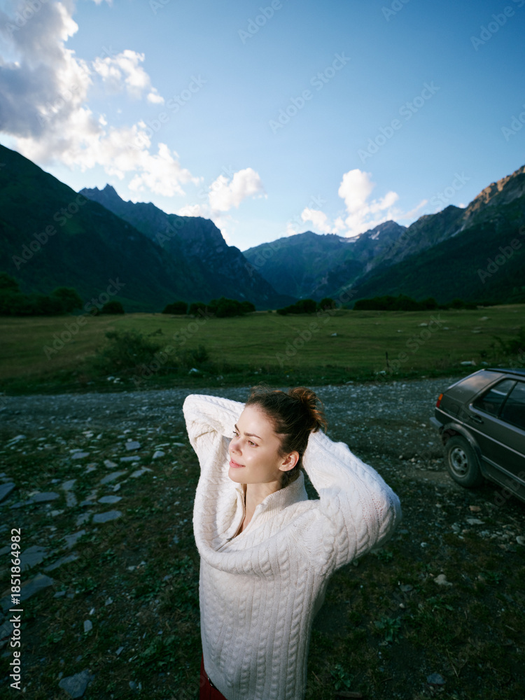 Fototapeta premium Woman wearing a chunky sweater in a mountain landscape, relaxing in a wide outdoor scene with green valley and blue sky, near a parked car during a peaceful countryside moment