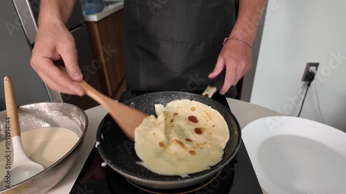 A man in an apron frying pancakes. Close-up. Flips a pancake with a wooden spatula on an induction hob