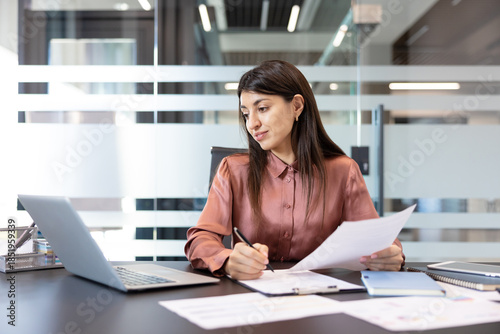 Businesswoman at office desk reviewing documents and taking notes on a clipboard while working on a laptop, focused on reports and planning in a modern corporate workspace