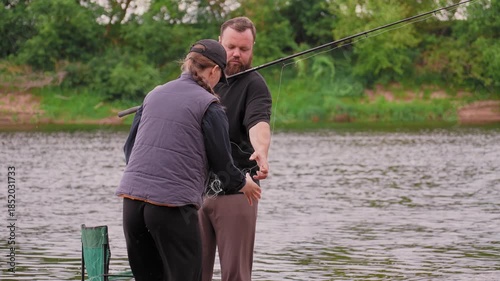 Middle-aged bearded man patiently teaching a young woman how to use a spinning reel on a fishing rod by the river, helping her untangle the fishing line for their joint hobby