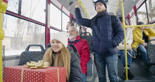 Side view of passengers traveling by bus. Small girl holding gift box, smiling, old man wearing red jacket, sitting, boy standing, handle holding. Concept of public transport.