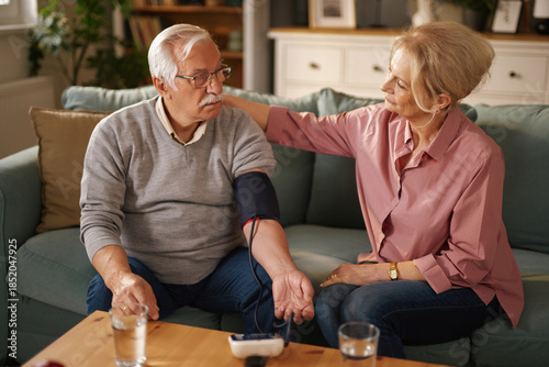Fotografie A blonde, older woman sits next to her gray-haired husband on a couch in their home and checks his blood pressure in the afternoon