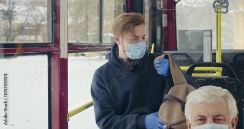 Front view of boy in mask and gloves sitting on bus. Passengers on bus, boy with earphones and rucksack, using smartphone, texting, putting in pocket. Concept of public transport.