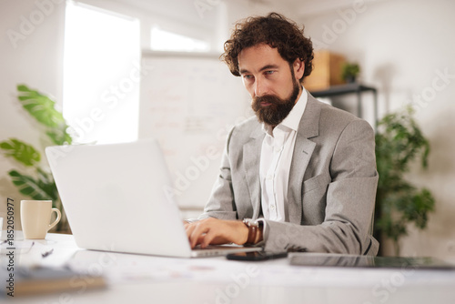 A man is sitting in an office chair at a desk. He is focused on his laptop while working. A cup is nearby and there are plants in the background.