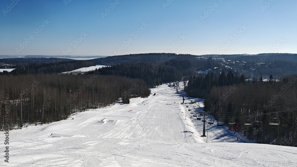 There are chairlifts and rope lifts on the slopes of the ski center. Skiers climb them. There are ski tracks in the snow. There is a forest around, and a village behind it. Sunny winter weather