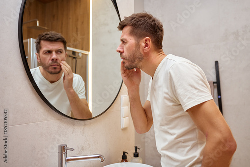 Man examines facial skin while looking at mirror in modern bathroom