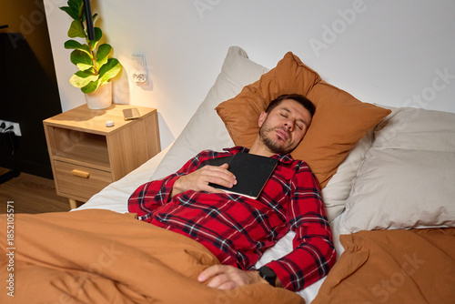 Fatigued man sleeping in bed holding notebook in bedroom