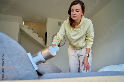 Woman using handheld vacuum cleaner to clean sofa in modern living room