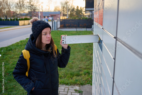 Woman wearing winter jacket and hat opening metal mailbox outside apartment building