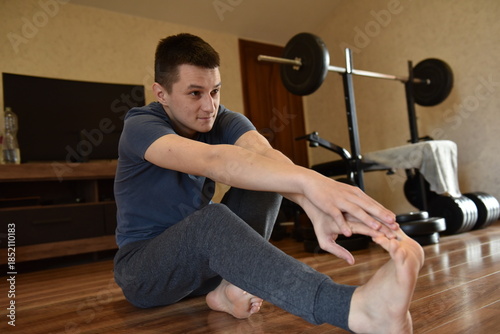 Young man stretching legs on wooden floor during home workout, flexibility exercise near weight bench in cozy apartment