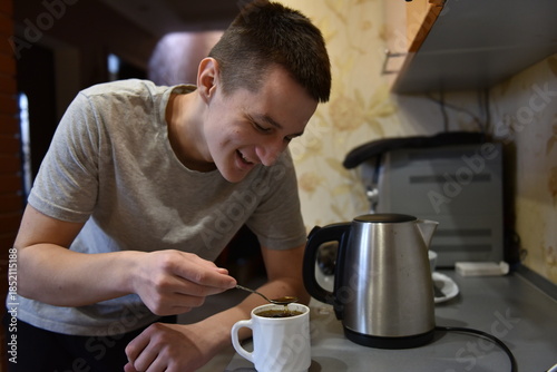 Young man smiling while making coffee at home kitchen, stirring hot drink with spoon near electric kettle in cozy morning routine