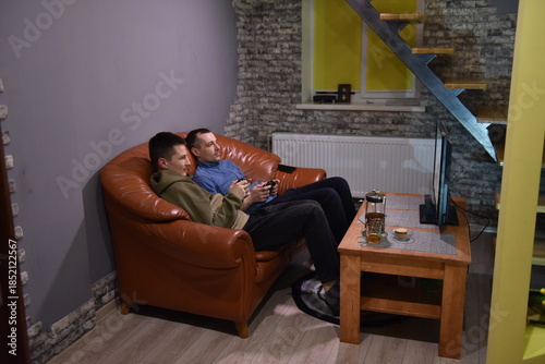Two men sitting on leather sofa playing video games at home, relaxed evening leisure time with console controllers and TV in modern living room