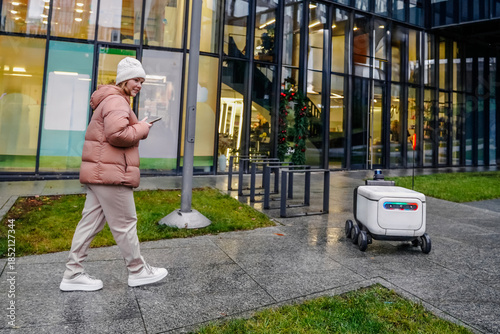 Delivery robot drives on wet pavement. Woman uses smartphone for autonomous courier tracking, real time navigation. Urban logistics service supports contactless package transport in rainy weather.