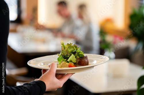 waiter presents a beautifully plated dish of meat medallions with a fresh green salad and garnish in a blurred, upscale restaurant setting