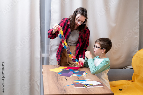 A young woman in a red plaid shirt makes a paper garland for Christmas with her son, who uses hearing aids. DIY Christmas home decor.
