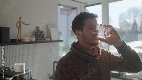 Adult man drinking fresh clean water from glass while standing in kitchen near window with sunlight. Person quenching thirst at home. Concept of hydration and healthy lifestyle