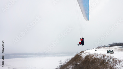 Winter paragliding. A paraglider takes off from a steep, snow-covered river slope. Below, the river is covered in ice.