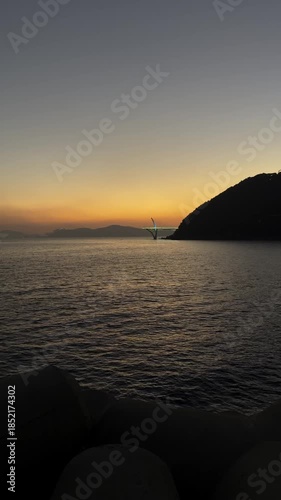 Beautiful calm ocean at dusk with an orange and yellow sunset. Gentle waves lapping against a rocky shore with distant islands and a bridge