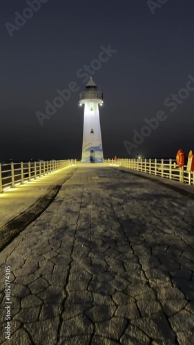 First-person perspective of someone walking on a stone pier toward a brightly lit white lighthouse against a dark night sky