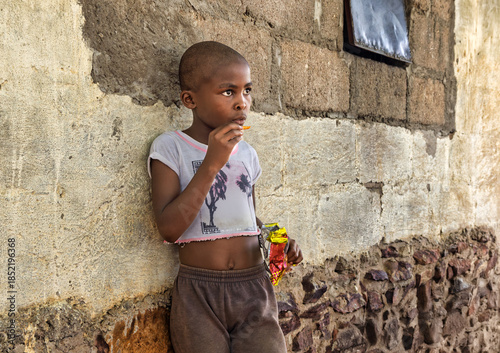 hungry single hungry african girl child in the village, bald shaved head, eating snacks, home next to the house wall,