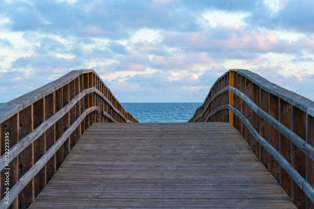 Fototapeta premium A wooden boardwalk curves gently toward the open ocean under a sky filled with soft clouds at sunset.