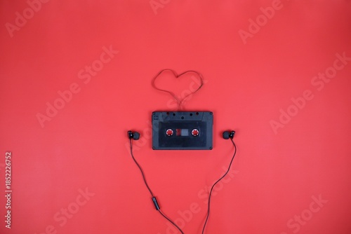 A vintage cassette tape sits on a red surface, with earbud cords forming a heart shape above it. A playful, nostalgic scene that conveys music, love, and creative connection.
