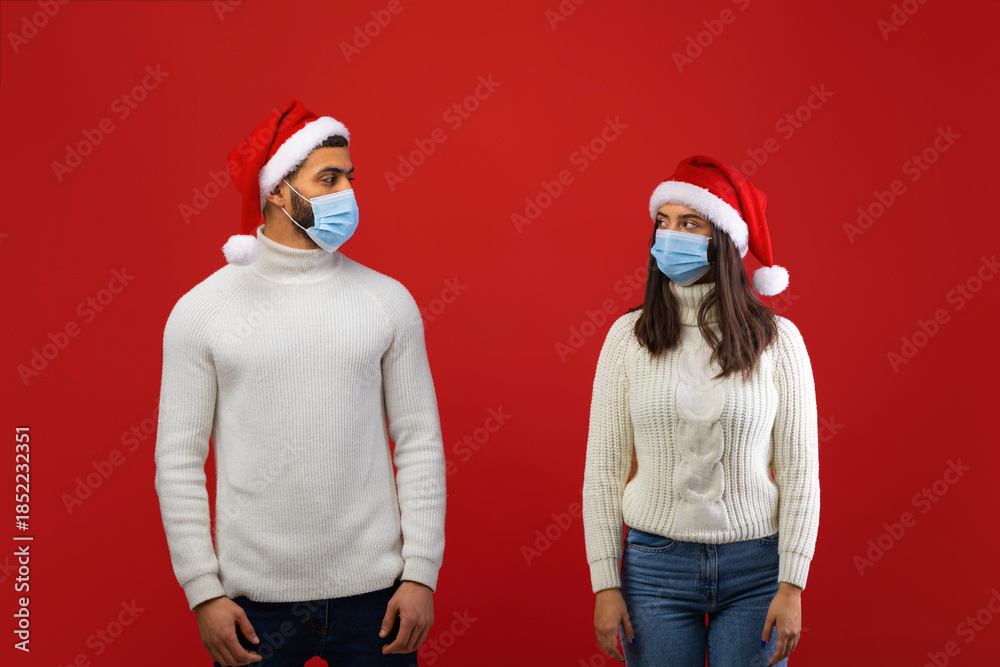 Fototapeta premium During the covid-19 lockdown, an Arab couple stands apart wearing Santa hats and protective masks, looking at each other. The red background highlights their festive spirit despite the situation.