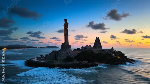 Dramatic sunset over a rocky sea island with a tall statue and distant shore.