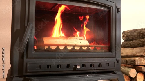 Burning wood stove. Cast iron stove with burning wood inside. Wood-burning stove in room with flames nside. Logs are stacked for fuel, warm glow fills space. It is winter, atmosphere feels inviting.