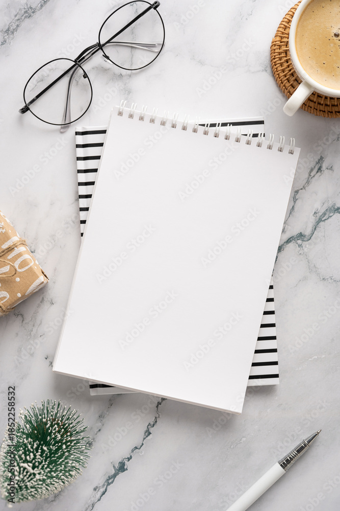 Obraz premium A stylish desk setup featuring a blank notebook on top of striped paper, glasses, a cup of coffee, a small wrapped gift, and seasonal decorations, perfect for holiday planning and creative work