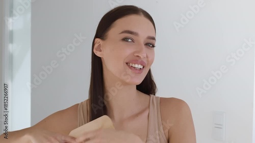 A woman with long hair is smiling as she brushes her hair in a bright, modern room. Natural light illuminates the space, creating a cheerful atmosphere.