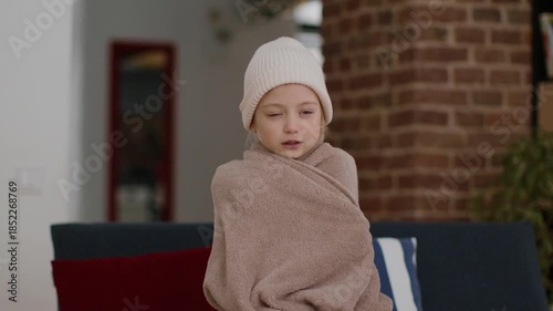 A young child sits comfortably wrapped in a soft blanket while wearing a warm hat. The cozy indoor setting features brick walls and colorful pillows, creating a peaceful winter atmosphere.