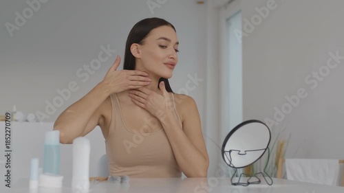 A woman sits at a clean, bright vanity. She gently applies skincare products while looking into a mirror. The setting is cozy and modern, with soft natural light coming through the window.