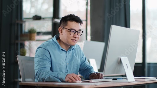 A man wearing glasses is focused on his work at a sleek desk in a contemporary office. Sunlight streams through large windows, creating a productive atmosphere.