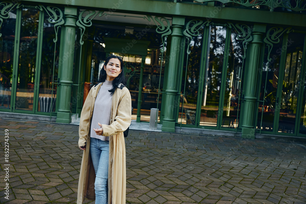 Naklejka premium Woman wearing a long beige coat stands outdoors in front of green glass doors, casual denim and sweater visible, framed by arches and a tiled courtyard beside architectural detail